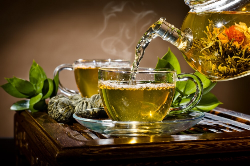 Uma chícara de vidro transparente transbordando com chá verde quente e fumegante, posicionada artisticamente ao lado de galhos frescos de folhas de chá verde, evocando um ambiente sereno e acolhedor para o consumo de chá.